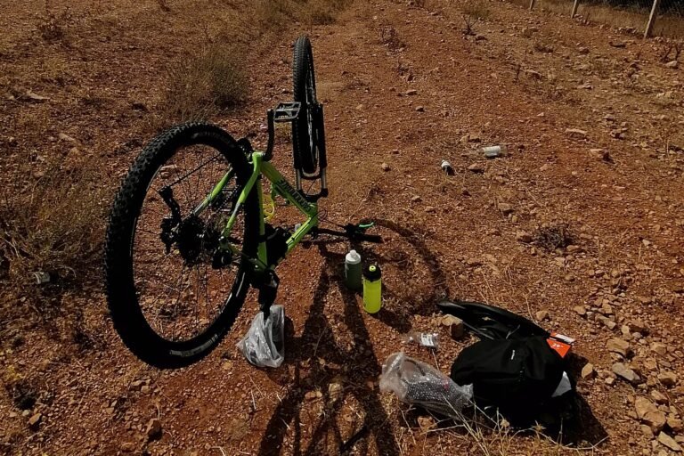 A mountain bike turned upside down at the roadside with its front wheel removed, tools, a bottle and a repair kit laid out on the ground beside it