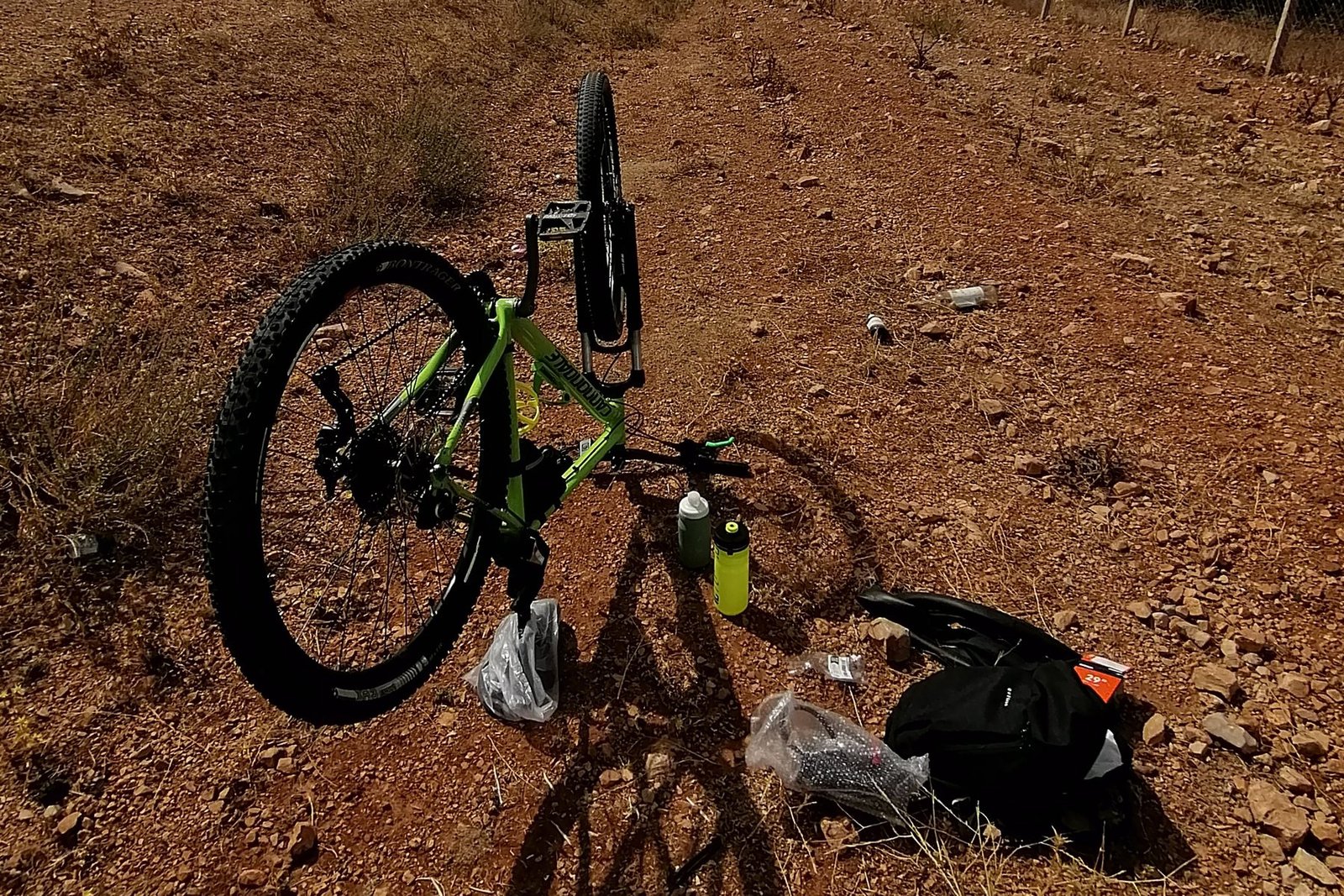 A mountain bike turned upside down at the roadside with its front wheel removed, tools, a bottle and a repair kit laid out on the ground beside it