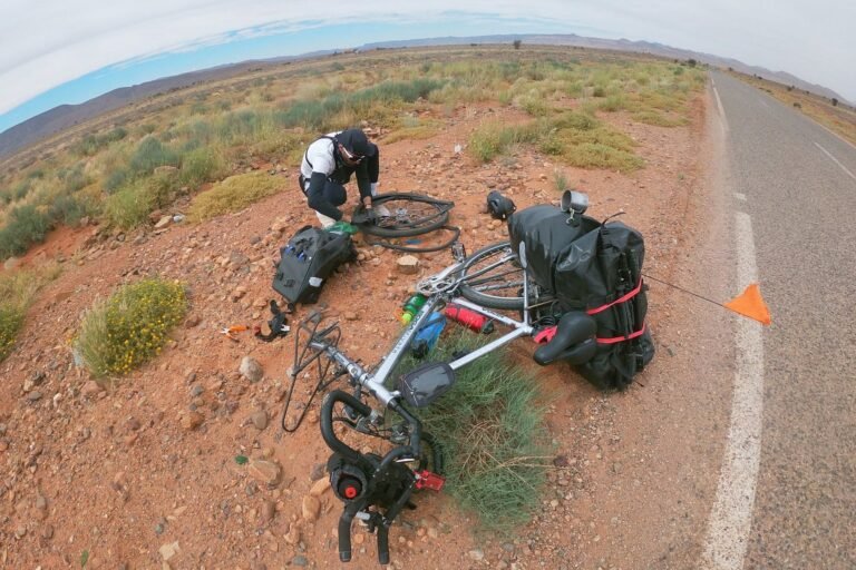A cyclist repairing a loaded touring bike at the roadside on a desert highway, with a warning triangle placed behind